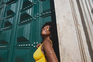 Siyah saçlı, sarı elbiseli güzel bir zenci kadın büyük yeşil kilise kapısına poz veriyor. Mutlu ve gülümsüyor