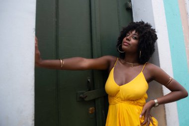 Afro saçlı, sarı elbiseli, yeşil kapıya dayanmış fotoğraf için poz veren güzel siyahi kadın portresi. Yolculukta mutlu insan.