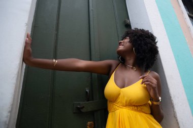 Afro saçlı, sarı elbiseli, yeşil kapıya dayanmış fotoğraf için poz veren güzel siyahi kadın portresi. Yolculukta mutlu insan.