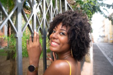 Eski demir bir çite dayanan sarı bir elbise giyen afro saçlı güzel siyah bir kadının portresi. Yolculuktaki mutlu insan..