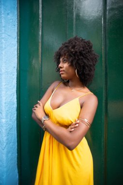 Afro saçlı, sarı elbiseli, kolları yeşil kapıya bağlı mutlu bir kadın. Yolculukta mutlu insan.