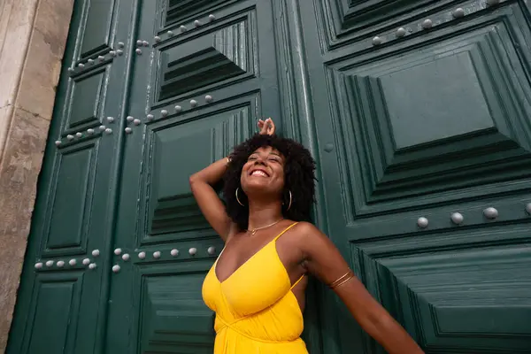 Sarı elbiseli, afro saçlı güzel siyah bir kadın büyük yeşil bir kapıya yaslanmış bir fotoğraf için poz veriyor. Mutlu ve gülümsüyor
