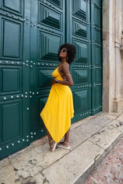 Sarı elbiseli, afro saçlı güzel siyah bir kadın büyük yeşil bir kapıya yaslanmış bir fotoğraf için poz veriyor. Mutlu ve gülümsüyor