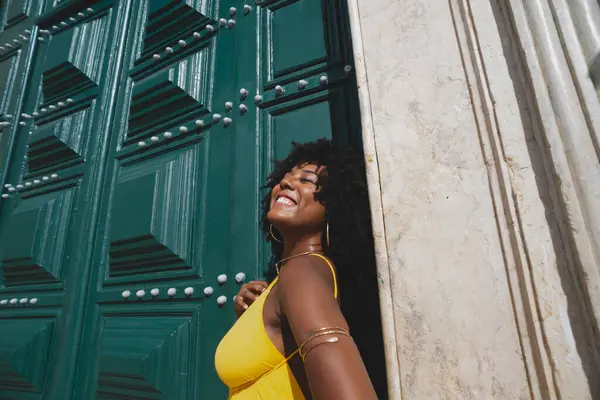 Siyah saçlı, sarı elbiseli güzel bir zenci kadın büyük yeşil kilise kapısına poz veriyor. Mutlu ve gülümsüyor