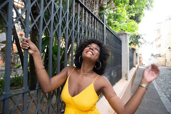Afro saçlı, sarı elbiseli, mutlu bir kadın eski demir bir çite yaslanmış kameraya poz veriyor. Yolculuktaki mutlu insan..