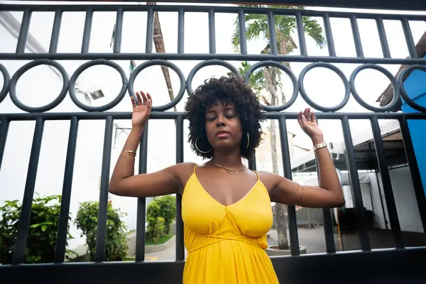 Demir bir çitin yanında sarı elbiseli, afro saçlı bir kadın. Kendinden emin ve mutlu biri.
