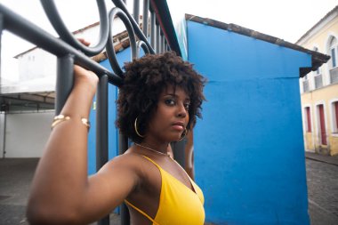 Demir bir çitin yanında sarı elbiseli, afro saçlı bir kadın. Kendinden emin ve mutlu biri.