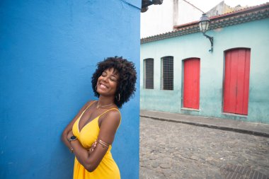 Sarı elbiseli, afro saçlı, mavi duvarda izole edilmiş ve arka planda evleri olan güzel bir kadın. Kendinden emin ve mutlu biri.