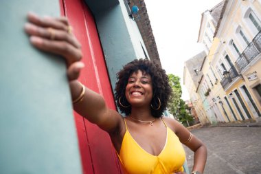 Sarı elbiseli, afro saçlı güzel bir kadın kırmızı kapının yanında fotoğraf çektirmek için poz veriyor. Kendinden emin ve mutlu biri. Brezilya