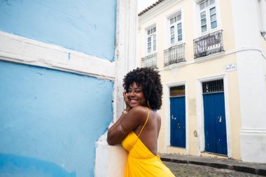 Sarı elbiseli ve afro saçlı güzel siyah kadın eski mavi bir eve yaslanıyor. Kendinden emin ve mutlu biri. Brezilya