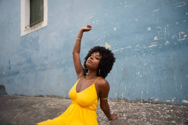 Sarı elbiseli ve afro saçlı güzel bir kadının portresi mavi duvara karşı eski caddenin kaldırımında oturuyor. Kendinden emin ve mutlu biri. Brezilya