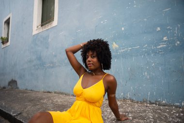 Sarı elbiseli ve afro saçlı güzel bir kadının portresi mavi duvara karşı eski caddenin kaldırımında oturuyor. Kendinden emin ve mutlu biri. Brezilya
