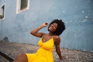 Sarı elbiseli ve afro saçlı güzel bir kadının portresi mavi duvara karşı eski caddenin kaldırımında oturuyor. Kendinden emin ve mutlu biri. Brezilya
