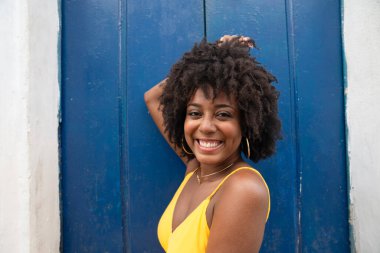 Siyah saçlı, sarı elbiseli, eski bir evin mavi kapısında oturan ve kameraya bakan genç bir kadının yakın plan portresi. Kendine güvenen biri. Brezilya