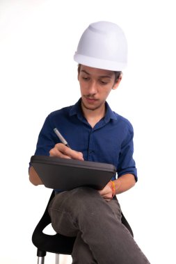 Genç mimar beyaz kask takıyor, oturuyor ve tabletle çalışıyor. Beyaz arka planda izole edilmiş. Güvenlik ekipmanları.