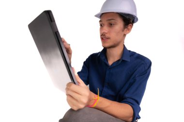 Genç mimar beyaz kask takıyor, oturuyor ve tabletle çalışıyor. Beyaz arka planda izole edilmiş. Güvenlik ekipmanları.