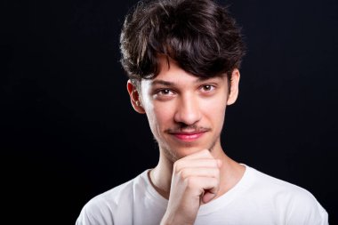 Beyaz gömlekli yakışıklı bir genç tek eliyle çenesine dokunuyor. Siyah arka planda izole edilmiş. Stüdyo çekimi