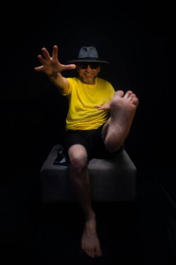 Olgun bir adam, 50 'li yaşlarda, oldukça çekingen, sakallı, güneş gözlüklü, şapkalı ve sarı gömlekli, puf minder üzerinde oturan bir fotoğraf için sakince poz veriyor. Brezilya