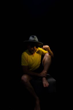 Olgun bir adam, 50 'li yaşlarda, sarı gömlek, güneş gözlüğü ve şapka takıyor, oturuyor, elleriyle poz veriyor. Şık bir insan. Brezilya