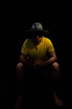 Olgun bir adam, 50 'li yaşlarda, sarı gömlek, güneş gözlüğü ve şapka takıyor, oturuyor, fotoğraf için poz veriyor. Sakin biri. Brezilya