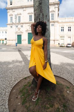 Afro saçlı, sarı elbiseli, fotoğraf için poz veren güzel siyah kadın kilise arka planında hindistan cevizi ağacı gövdesine yaslanıyor. Mutlu gülümseyen kişi