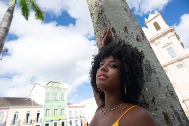 Afro saçlı, sarı elbiseli, fotoğraf için poz veren güzel siyah kadın kilise arka planında hindistan cevizi ağacı gövdesine yaslanıyor. Mutlu gülümseyen kişi