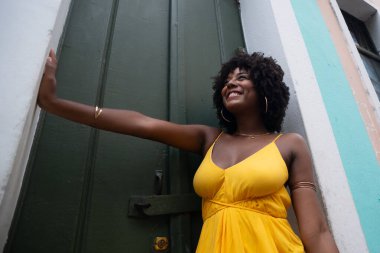 Afro saçlı, sarı elbiseli, yeşil kapıya dayanmış fotoğraf için poz veren güzel siyahi kadın portresi. Yolculukta mutlu insan.