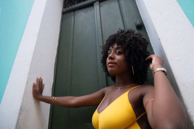 Afro saçlı, sarı elbiseli, yeşil kapıya dayanmış fotoğraf için poz veren güzel siyahi kadın portresi. Yolculukta mutlu insan.