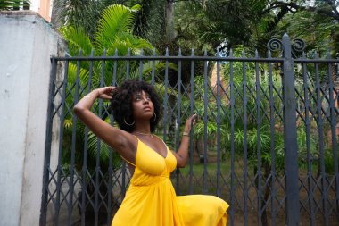 Eski demir bir çite dayanan sarı bir elbise giyen afro saçlı güzel siyah bir kadının portresi. Yolculuktaki mutlu insan..