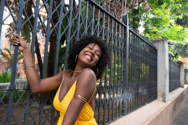 Afro saçlı, sarı elbiseli, mutlu bir kadın eski demir bir çite yaslanmış kameraya poz veriyor. Yolculuktaki mutlu insan..