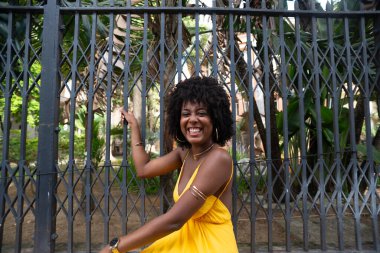 Afro saçlı, sarı elbiseli, mutlu bir kadın eski demir bir çite yaslanmış kameraya poz veriyor. Yolculuktaki mutlu insan..