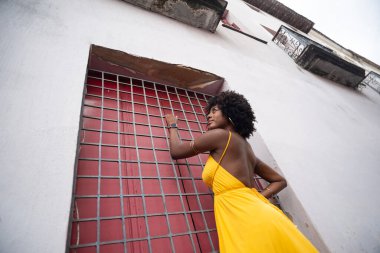 Sarı elbiseli, afro saçlı, demir parmaklık tutan, kırmızı kapılı, yana bakan bir kadın. Kendinden emin ve mutlu biri.