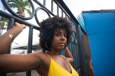 Demir bir çitin yanında sarı elbiseli, afro saçlı bir kadın. Kendinden emin ve mutlu biri.