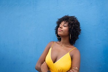 Sarı elbiseli, afro saçlı, mavi renkli bir duvarda izole edilmiş güzel bir kadın. Kendinden emin ve mutlu biri.