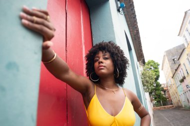 Sarı elbiseli, afro saçlı güzel bir kadın kırmızı kapının yanında fotoğraf çektirmek için poz veriyor. Kendinden emin ve mutlu biri. Brezilya