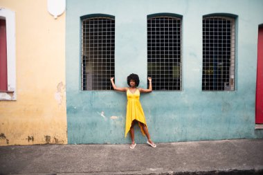Sarı elbiseli ve afro saçlı güzel bir kadın pencereler arasındaki evin duvarına yaslanmış bir fotoğraf için poz veriyor. Kendinden emin ve mutlu biri. Brezilya