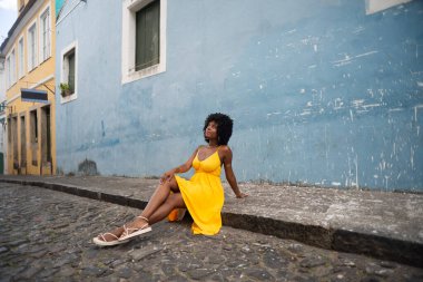 Sarı elbiseli ve afro saçlı güzel bir kadının portresi mavi duvara karşı eski caddenin kaldırımında oturuyor. Kendinden emin ve mutlu biri. Brezilya
