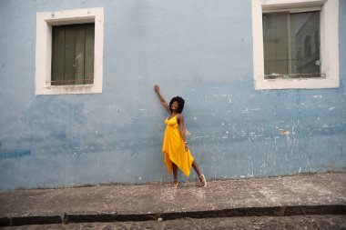 Sarı elbiseli ve afro saçlı güzel siyah bir kadın bir evin pencereleri arasında fotoğraf çekmek için poz veriyor. Kendinden emin ve mutlu biri. Brezilya