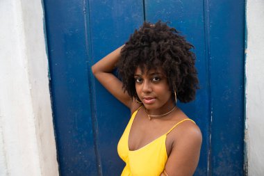 Siyah saçlı, sarı elbiseli, eski bir evin mavi kapısında oturan ve kameraya bakan genç bir kadının yakın plan portresi. Kendine güvenen biri. Brezilya