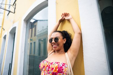 Örgülü, renkli elbiseli, kolları havada ve güneş gözlüğü takan beyaz bir kadın. Seyahat eden biri. Brezilya