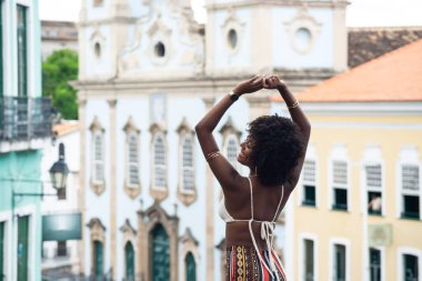Siyah saçlı genç bir kadın, kameraya döndü, kollarını kaldırdı. Arka plandaki eski evlere karşı. Mutlu ve güçlü biri. Brezilya