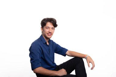 Siyah saçlı, mavi gömlekli, oturan, gülümseyen ve kameraya bakan yakışıklı bir genç. Beyaz arka planda izole edilmiş. Kendine güvenen biri. Profesyonel konsept.