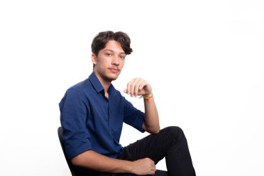 Siyah saçlı, mavi gömlekli, oturan ve kameraya ciddi ciddi bakan genç ve yakışıklı bir öğrenci. Beyaz arka planda izole edilmiş. Kendine güvenen biri. Profesyonel konsept.