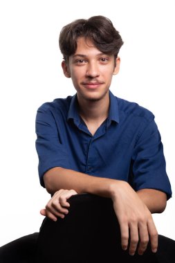 Siyah saçlı, mavi gömlekli, oturan ve kameraya ciddi ciddi bakan genç ve yakışıklı bir öğrenci. Beyaz arka planda izole edilmiş. Kendine güvenen biri. Profesyonel konsept.