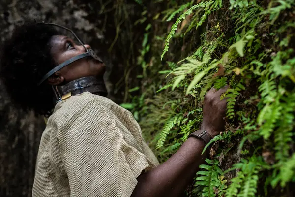 Ağzı ve boynu demir maskeli bir kadın yeşil bitkilerle dolu bir duvarın yanına bakıyor. Brezilya 'da kölelik. Köle Anastacia 'nın temsilcisi.