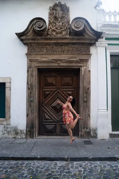 Renkli elbiseler giyen, zıplayan ve kameraya poz veren, büyük, eski ahşap bir kapıya bakan kadın. Seyahat eden biri. Brezilya