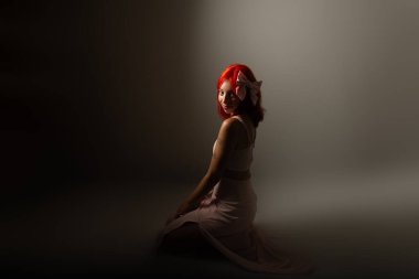 Pembe elbiseli genç, kızıl saçlı kadın yere çömelmiş, karanlık arka planda fotoğraf için poz veriyor. Stüdyo resmi