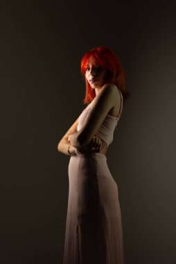 Kolları çapraz duran, koyu arkaplanda fotoğraf çektirmek için poz veren güzel, kızıl saçlı bir kız. Stüdyo resmi