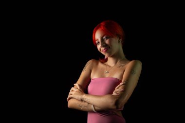 Genç, güzel, kızıl saçlı, pembe elbiseli bir kadın koyu arkaplanda fotoğraf için poz veriyor. Stüdyo resmi.