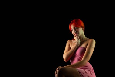 Güzel, genç, kızıl saçlı bir kadının portresi, oturan, pembe elbiseli, koyu arkaplanda fotoğraf için poz veren. Stüdyo resmi.